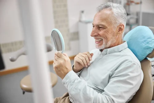 contented elderly man sitting dental chair checking out his new teeth while looking mirror